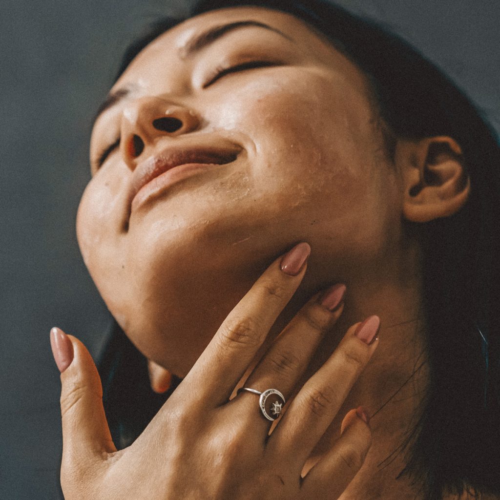 Eine Frau mit langem, dunklem Haar hat den Kopf in den Nacken gelegt, die Augen geschlossen und l&auml;chelt sanft. Sie ber&uuml;hrt mit der linken Hand ihren Hals und tr&auml;gt einen silbernen Ring mit einem kleinen Blumenmuster. Der Hintergrund ist dunkel und unterstreicht ihren heiteren Gesichtsausdruck.