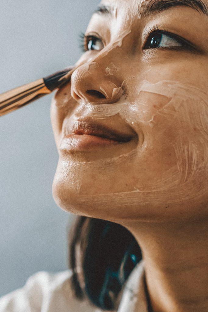 Eine Person l&auml;chelt, w&auml;hrend sie mit einem Pinsel eine Gesichtsmaske auftr&auml;gt. Die Maske wird auf der Wange und um die Augenpartie verteilt. Der Hintergrund ist sanft verschwommen, wodurch der Ausdruck und die glatte Textur der Maske hervorgehoben werden.